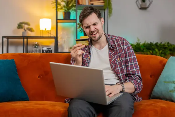 Young sad man guy in plaid shirt sits on couch using laptop and credit card frowns trying to make payment through internet feels angry due lack of money bankruptcy. Caucasian victim of fraud concept
