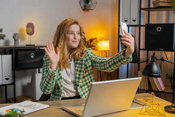 Hello. Happy businesswoman waves hand hi gesture greeting welcomes someone on smartphone, online selfie webinar, remote distance conversation. Freelance sitting at desk using laptop in home office