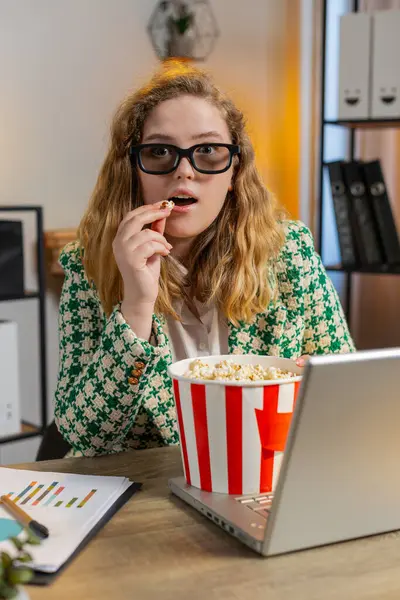 Businesswoman freelancer taking break from work wearing 3D glasses eating popcorn and watching movie on laptop. Smiling blond girl sitting at home office desk having snacks during break. Vertical