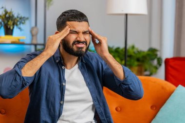 Hasta Hintli adam baş ağrısı problemini tedavi etmek için tapınakları ovuyor, tansiyon ve migren stresinden muzdarip evde acı içinde kıvranıyor yüksek tansiyon. Oturma odasındaki kanepede oturan üzgün İspanyol adam..