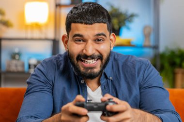 Neşeli mutlu Hintli adam oyun kumandası kullanıyor. Oturma odasında kanepede oturup oyun oynuyor. İspanyol yumruk sallayan adam hafta sonu evde online maç kazanmanın keyfini çıkarıyor..