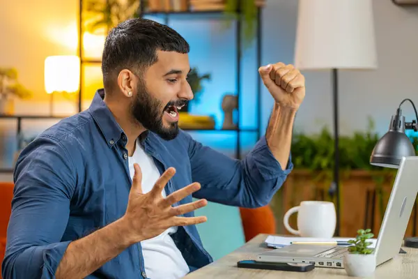 Happy amazed Indian man working on laptop shocked by sudden victory, celebrate game winning, goal achievement, good career news. Excited Arabian freelancer at home office workplace table desk.