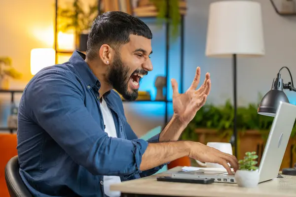 Angry furious Indian man freelancer using laptop while working at home office having nervous breakdown at work, stress management, mental distress problems, losing temper, reaction on failure.
