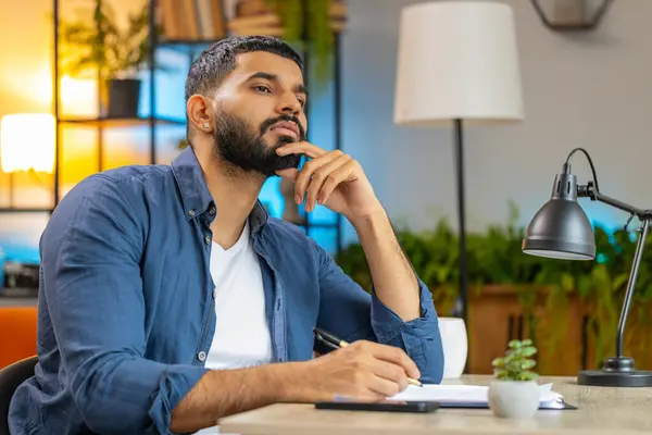 Indian bearded man freelancer working with documents from home office, analyzing information, reading business papers prepare financial report. Young Arabian guy accountant analyzing graph documents.
