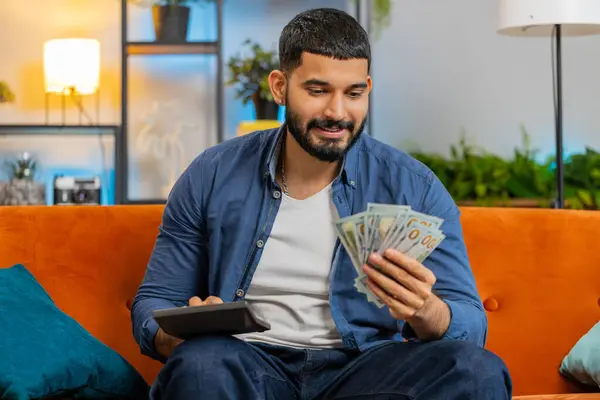 Planning budget. Rich happy Indian man using calculator counting money cash, calculate domestic bills at home. Hispanic guy satisfied of income earnings win saves money for planned vacation gifts.
