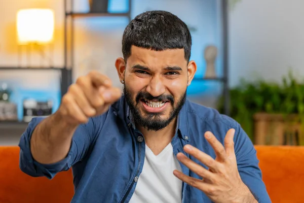 Hey you. Young upset bearded Indian man sitting on sofa looking with rage aggression, scolding protest, fighting to camera, displeasure, quarrel. Arabian guy accusing for mistake at home apartment.