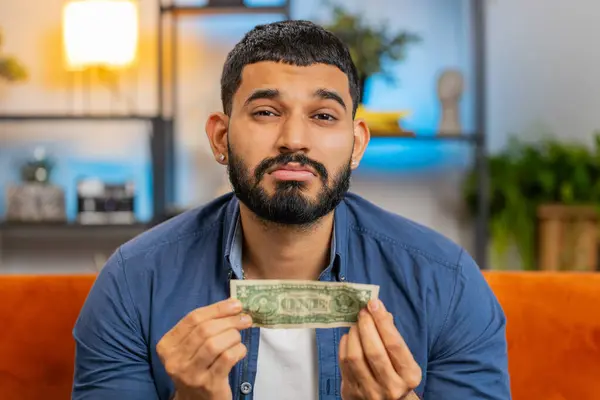Portrait of poor Indian Arabian man upset over insufficient amount of money showing one dollar banknote. Financial crisis. Bankruptcy. Poverty destitution. Upset Hispanic guy sitting on sofa at home