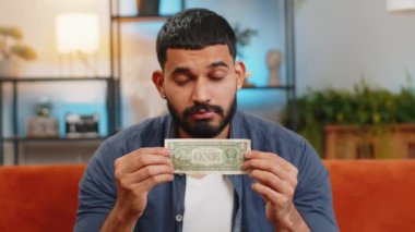 Zavallı Hintli adamın portresi yetersiz miktarda para için üzülüyor ve bir dolarlık banknot gösteriyor. Finansal kriz. İflas. Yoksulluk ve yoksulluk. Evdeki kanepede oturan üzgün İspanyol adam..