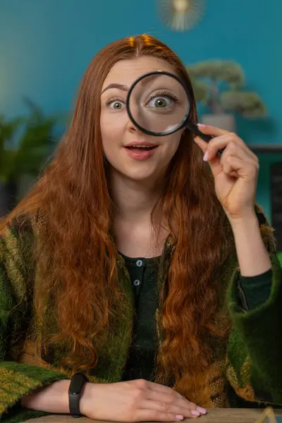 Investigator excited amazed Caucasian woman holding magnifying glass near face, looking into camera with big zoomed funny eye, wow, searching, analyzing. Girl at home office table workplace. Vertical