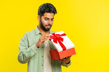 Mutlu Hintli genç adam kırmızı kurdeleli doğum günü hediyesi kutusunu açıyor. Tatil sürprizi konsepti. Gülümseyen Arap sakallı adam sarı stüdyo arka planında izole edilmiş büyük bir kutlama hediyesi aldı.