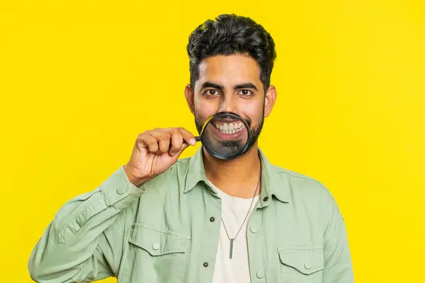 Happy Indian young man holding magnifier glass on teeth, looking at camera, showing funny smiling silly face mouth. Health care, hygiene, stomatology. Arabian guy isolated on studio yellow background