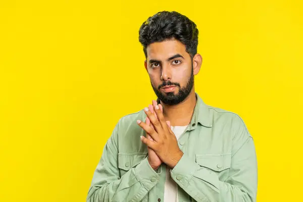 Sneaky cunning Indian young man with tricky face gesticulating and scheming evil plan, thinking over devious villain idea, cheats, jokes, pranks. Arabian Hindu guy isolated on yellow studio background