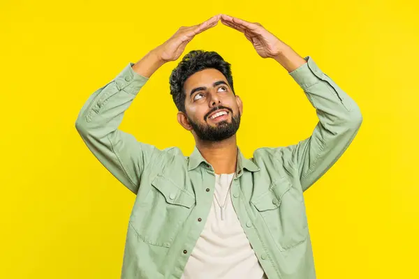 House roof about head. Indian young man playing childish catching up game, feeling in safe making roof above head with hands, insurance, security service. Arabian guy isolated on yellow background