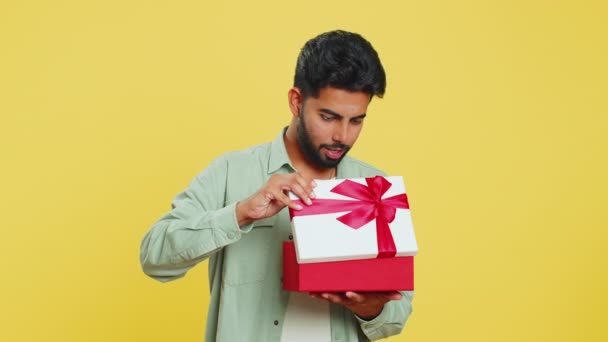 幸せなインドの若者は赤いリボンで誕生日のギフトボックスを開きます 休日はサプライズコンセプト 笑顔のアラビアひげの男は黄色いスタジオの背景で孤立した祝う偉大なラップされたプレゼントを受け取ります — ストック動画