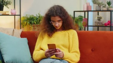 Mutlu çocuk kız cep telefonu daktilo kullanıyor. İnternette yazı yazıyor. Büyük bir sürpriz, güzel bir pozitif haber. Oturma odasında oturan genç kız kanepede oturuyor.