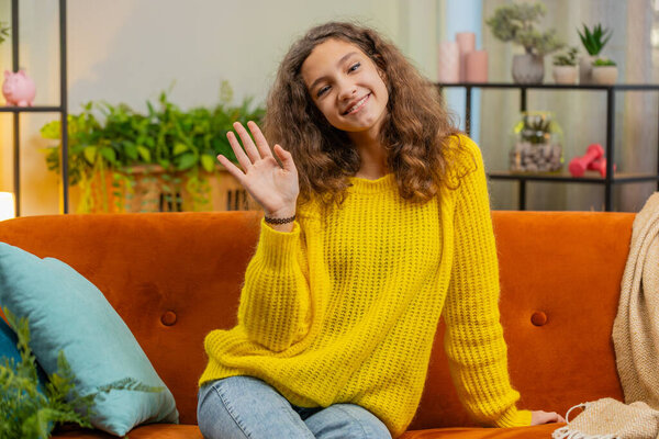 Young child girl smiling friendly at camera and waving hands gesturing hello, hi, greeting or goodbye, welcoming with hospitable expression. Teenager Caucasian kid at home in living room sits on sofa