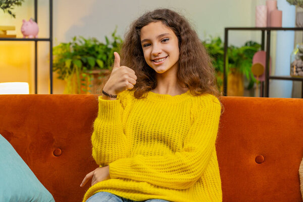 Like. Happy young Caucasian child girl looking approvingly at camera showing thumbs up like sign positive something good, positive feedback. Female teenager kid at home in living room sits on sofa