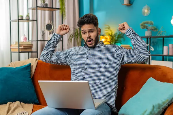 Surprised happy Indian man using laptop computer, receive mail good news message shocked by sudden victory celebrate lottery jackpot win purchases online shopping play game at home apartment on sofa