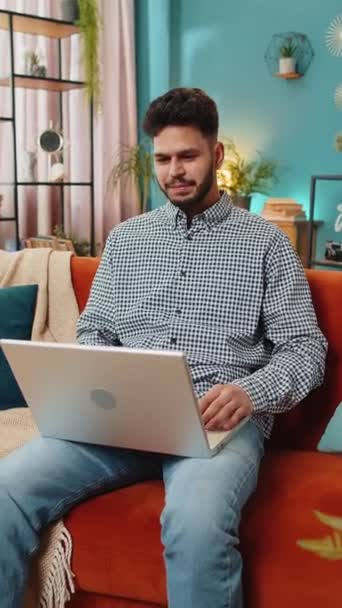 リビングで仕事を終えた後 ソファの閉じたノートパソコンに座っているインド人男性 アラビア人のフリーランサーは ホームソファーでオンラインリモートの仕事をしています ノートブックコンピュータでEラーニングブラウジングインターネット バーティカル — ストック動画