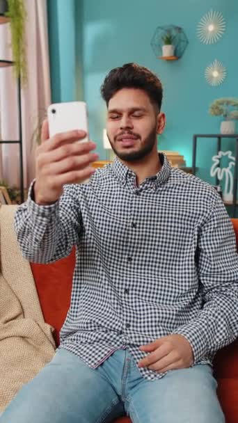 ハッピーインドのアラビア人男性ブロガーは スマートフォンでセルフィーを取ります ソーシャルメディアのためのストーリーを記録購読者とオンラインビデオ通話 ホームソファーに座っているヒスパニック系の男 バーティカル — ストック動画