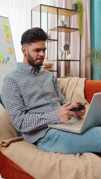 エキサイティングなインドのアラビアの男は 自宅のソファーソファーでラップトップの過ごし時間にビデオゲームをプレイしています ハッピーヒスパニックフリーランサーの男は コンピュータのPc上でジョイスティックコントローラでビデオゲームをプレイしています バーティカル — ストック動画