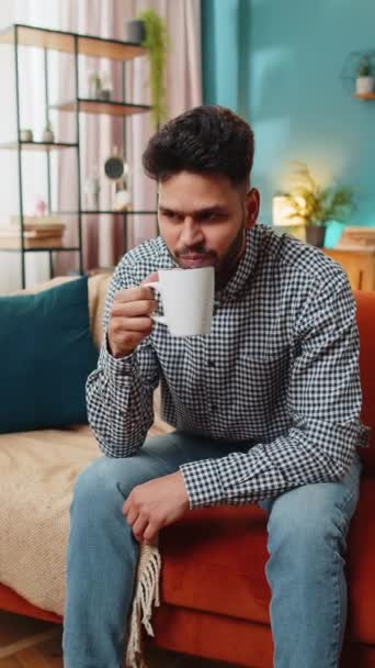 暖かいコーヒーやハーブティーを飲むインドの若い男性が 朝の自宅のリビングルームのソファーに座っています 一人でリラックスできるスペイン人男性 リラックス — ストック動画