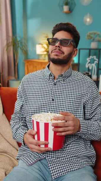 インド人男性フリーランサーは テレビで映画のコンテンツを見ているポップコーンを食べる3Dメガネを着て仕事から休憩します 笑顔のアラビア人の男は 週末に軽食を持っている家庭のソファーに座っています バーティカル — ストック動画