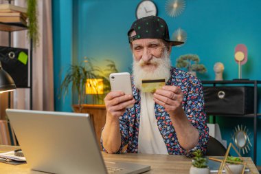 Merkez ofisteki online alışveriş ödemeleri için banka kartı ve dizüstü bilgisayar kullanan Kafkasyalı kıdemli bir adam. İş anlaşması. Gülümseyen şık büyükbaba satın alımları netbook E-bankacılık uygulama hizmeti ile.