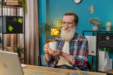 Üst düzey yaşlı işadamı dizüstü bilgisayarda çalışıyor sıcak kahve içiyor ve sabahları çalışma odasındaki masada oturuyor. Büyükbaba serbest çalışıyor. Dinlenmenin tadını çıkarıyor.