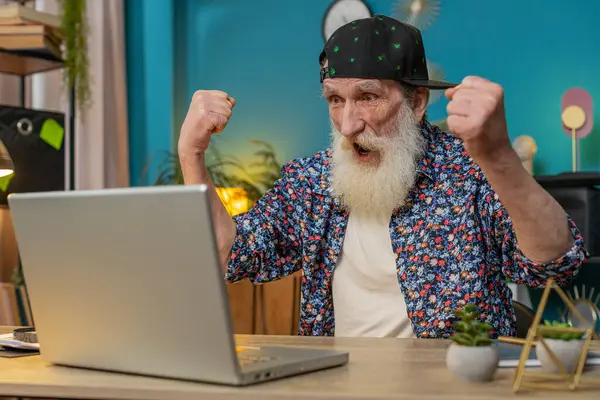 Happy senior old man winner in trendy shirt and cap at home office with laptop scream in triumph gesture celebrate success win money in lottery. Excited elderly mature grandfather get online good news