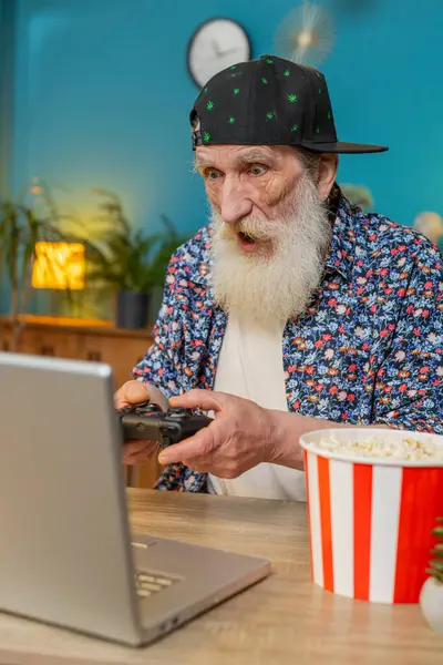 Excited senior old man celebrating victory in video game on laptop clenching fists, spending leisure time at home office desk. Happy stylish grandpa playing video games holding controller. Vertical