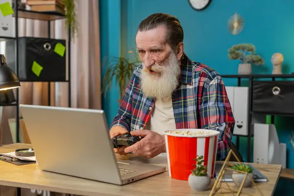 Excited Caucasian senior man celebrating victory in video game on laptop, spending leisure time at home office desk. Happy freelancer elderly mature old grandpa playing video games holding controller.