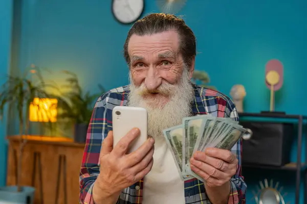 Rich Caucasian senior man counting money while having video call on smartphone sitting in home office. Male bearded freelancer old elderly mature grandpa satisfied of income earnings saves money