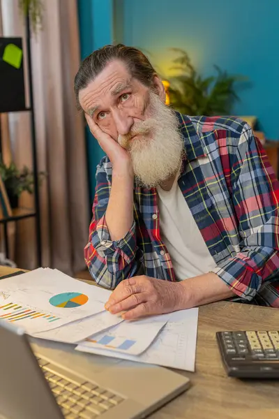 Tired exhausted senior man freelancer analyzing paperwork graphs, accounting payments and calculating monthly expenses bank bills at home office desk. Upset mature grandpa working remote job at table.