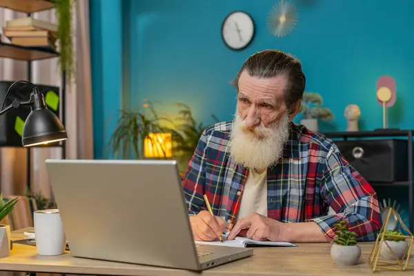 Caucasian senior businessman working on laptop computer online, writing notes in planner notebook at office workplace desk. Professional freelancer grandpa. Remote education, study, learning lessons.