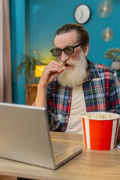 Senior man freelancer taking break from work wearing 3D glasses eating popcorn and watching movie on laptop. Laughing grandfather sitting at home office desk having snacks during work break. Vertical