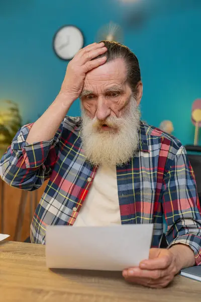 Shocked senior man freelancer analyzing paperwork graphs, charts, accounting payments and calculating monthly expenses bank bills at home office desk. Upset sad grandfather working remote job at desk