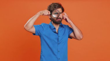 Araştırmacı araştırmacı bilim adamı elinde büyüteçle kameraya bakarken zumlanmış komik gözlerle analiz yapıyor. Genç Arap Orta Doğulu adam turuncu arka planda izole edilmiş.