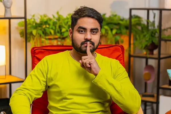Shh be quiet please. Indian Hindu confident man presses index finger to lips makes silence hush gesture sign do not tells gossip secret. Arabian guy at home apartment living room sitting on chair