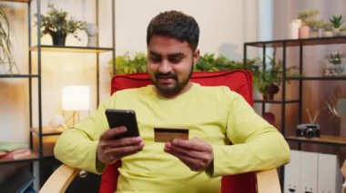 Hintli bir adam parayı transfer ederken kredi kartı ve akıllı telefon kullanıyor. İnternetten nakitsiz alışveriş yapıyor. Ev içinde yemek siparişi veriyor. Mutlu Arap adam sandalyede oturuyor.