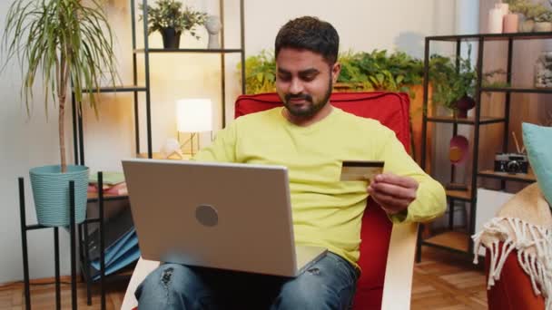 インドの男性は クレジットバンクのプラスチックカードとラップトップコンピュータを使用して お金を転送しながら オンラインショッピングを購入し 現代のホームルームアパートでインターネット上の食品配達を注文します 椅子に座っている男 — ストック動画