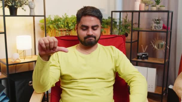 気に入らない 不幸な若いアラビア人男性は サインジェスチャーの親指を示し 不満を表明し 家のアパートで不満の悪い仕事を屋内で表現しました 椅子の部屋で不満なインディアン男 — ストック動画