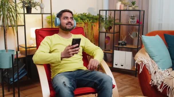 自宅で椅子でダンスするワイヤレスヘッドフォンで ハッピーリラックスしたインドのアラビア人男性は スマートフォンの楽しみの中で好きなエネルギッシュディスコポップミュージックを聞くことを選択します 週末の活動 — ストック動画