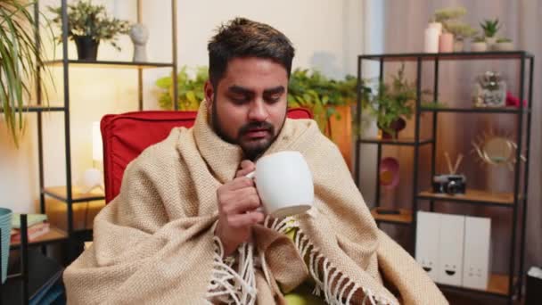 病気のインディアン男性は ソファーで冷たい風邪から一人で座って 熱くないアパートで熱いお茶を飲んで 負債を加えずに座っています 不健康なアラビア人の男が椅子に座って不快感を和らげようとします — ストック動画