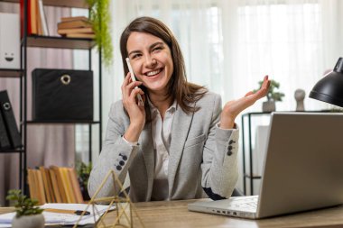 Gülümseyen beyaz iş kadını operatörü, müşterisiyle akıllı telefondan iletişim kuruyor, kağıt belgelerle notlar alıyor, ofis masasında dizüstü bilgisayarla çalışıyor. İnternetteki uzaklık işi kavramı.