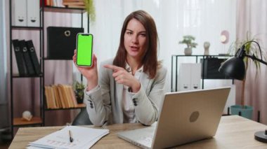 Beyaz iş kadınının elinde yeşil ekran krom tuşlu akıllı telefon olması iyi bir uygulama tanıtım teklifi sunuyor. Ofis masasında kameraya bakan serbest çalışan kadın..