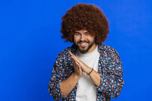 Sneaky cunning young man with Afro hairstyle wig with tricky face gesticulating and scheming evil plan, thinking over devious villain idea, cheats, jokes, pranks. Guy isolated on blue background