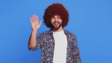Afro saç stili peruklu bir adam kameraya gülümseyerek el sallayarak selam veriyor ya da davetkar bir yüz ifadesiyle veda ediyor. Mavi stüdyo arka planında izole edilmiş şık bir adam.