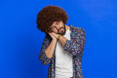 Yorgun, genç, uykulu, Afro saç stili peruklu, dikkatsiz, uykusuzluktan ağzı açık, uykusuz, yorgun ve yorgun bir adam. Stüdyo mavi arka planda izole adam