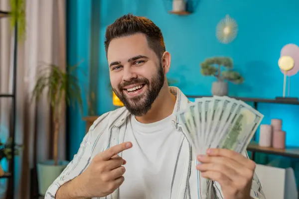 Young happy businessman guy holding fan of cash money dollar banknotes showing pointing on banknotes, celebrate, success career, online income, wealth at home office. Remote distant working